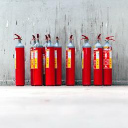 Eight red fire extinguishers aligned in front of a gray concrete wall, resembling a choir standing in formation.