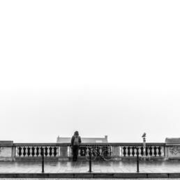 A lone person stands with their back turned on a foggy urban terrace, next to a bicycle, facing a balustrade overlooking a barely visible cityscape.
