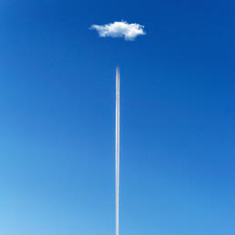 A lone airplane leaves a vertical contrail directly beneath a single fluffy cloud in a clear blue sky.