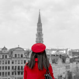A woman in a vivid red coat and beret stands with her back to the viewer, facing the gray-toned skyline of Brussels, with the spire of the Town Hall rising in the distance.