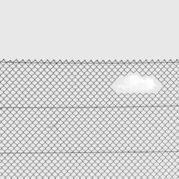 A chain-link fence with a cloud-shaped hole in it, allowing a white cloud in the sky to be visible through the gap.
