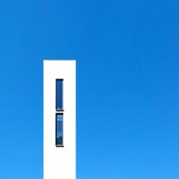 A tall, narrow white building facade with two vertically aligned windows, set against a clear blue sky.