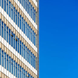 A modernist office building photographed from the side, its repetitive glass windows and beige facade contrast with a perfectly clear, deep blue sky.