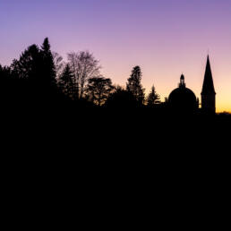 Silhouettes of trees and a church dome with spire against a purple and orange sunset sky.