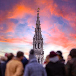 Crowd of people in the foreground looking at a tall Gothic tower spire against a colorful sunset sky.