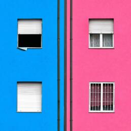 A building façade divided vertically: the left side painted bright blue with two windows, the right side painted bright pink with two windows. Black drainage pipes run vertically down the middle, separating the two color fields.