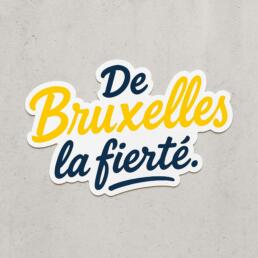 Sticker-style graphic with the phrase ‘De Bruxelles la fierté’ in cursive yellow and blue lettering on a grey background.